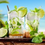 Mojito analcolico: la ricetta del cocktail fresco del virgin mojito 1 con lo sfondo di una spiaggia tropicale con le palme, tavolo di legno con tagliere e sopra 2 bicchieri con mojito analcolico con lime e foglie di menta e ghiacchio