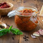Pesto di pomodori secchi, la salsa veloce dal gusto mediterraneo 2 Vasetto di pesto di pomodori secchi con gli ingredienti principali sul tavolo: basilico, aglio, mandorle e parmigiano.