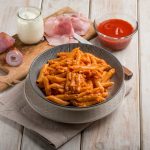 Penne al baffo: un primo piatto facilissimo e delizioso 1 su tavolo di legno piatto fondo con penne al baffo e accanto tagliere con gli ingredienti cipolla, panna, prosciutto cotto e sugo di pomodori