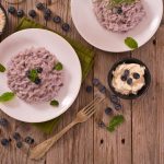 Risotto ai mirtilli neri: ricetta facile con mirtilli freschi o surgelati 1 Risotto ai mirtilli servito su piatti bianchi con decorazione di foglie di menta, affiancato da ciotoline con riso, mirtilli freschi e crema al formaggio, su tavolo rustico in legno.