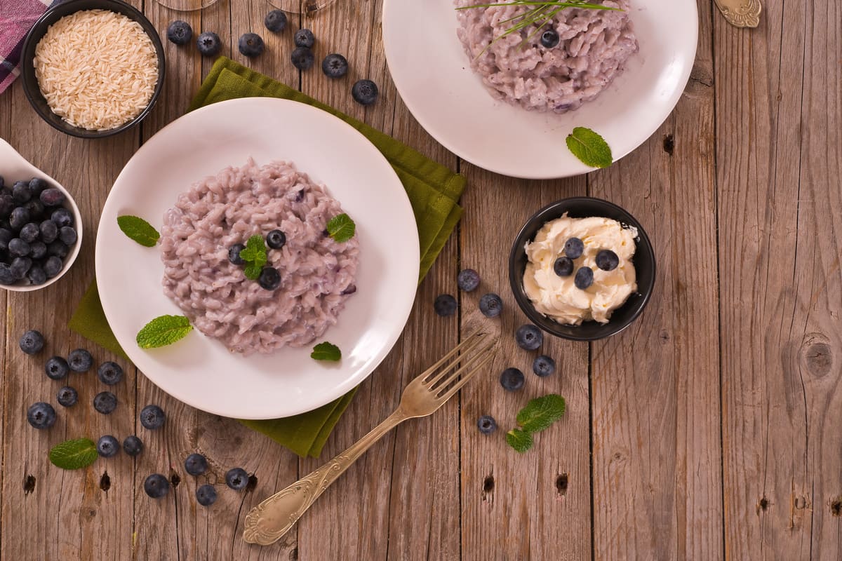 Ricette con i mirtilli: idee colorate e gustose 15 Risotto ai mirtilli servito su piatti bianchi con decorazione di foglie di menta, affiancato da ciotoline con riso, mirtilli freschi e crema al formaggio, su tavolo rustico in legno.