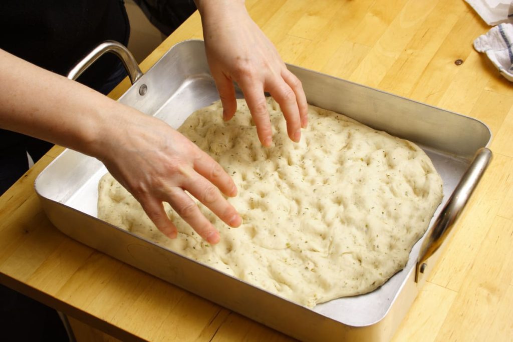 Schiacciata toscana: ricetta originale e varianti con lievito e Bimby 1 In primo piano delle mani che stendono l’impasto della schiacciata in una teglia rettangolare. La pasta, già lievitata e aromatizzata con erbe, viene pressata con le dita per creare la tipica superficie irregolare prima di essere infornata.