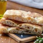 Schiacciata toscana: ricetta originale e varianti con lievito e Bimby 2 Tre fette di schiacciata sovrapposte su un tagliere, con crosta dorata e interno soffice e ben alveolato, accompagnate da sale grosso e olio extravergine di oliva in secondo piano.