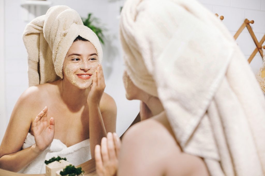 Scrub viso: cos’è, benefici, come esfoliare la pelle e migliori prodotti secondo le recensioni Amazon 1 donna allo specchio con asciugamano nei capelli e attorno al corpo. e' sorridente e sta applicando uno scrub viso