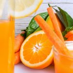 Succo ACE fatto in casa con il frullatore o l'estrattore 1 primo piano di un bicchiere di succo ACE con una fette di limone sul bicchiere e sulla destra gli ingredienti per fare un succo pieno di vitamine: arancia, carote e erbe aromatiche