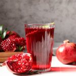 Succo di melograno: la ricetta per farlo in casa, benefici e come conservarlo 3 Un bicchiere di succo di melograno dal colore rosso intenso è posizionato al centro, circondato da frutti interi e tagliati che mostrano i chicchi lucidi.