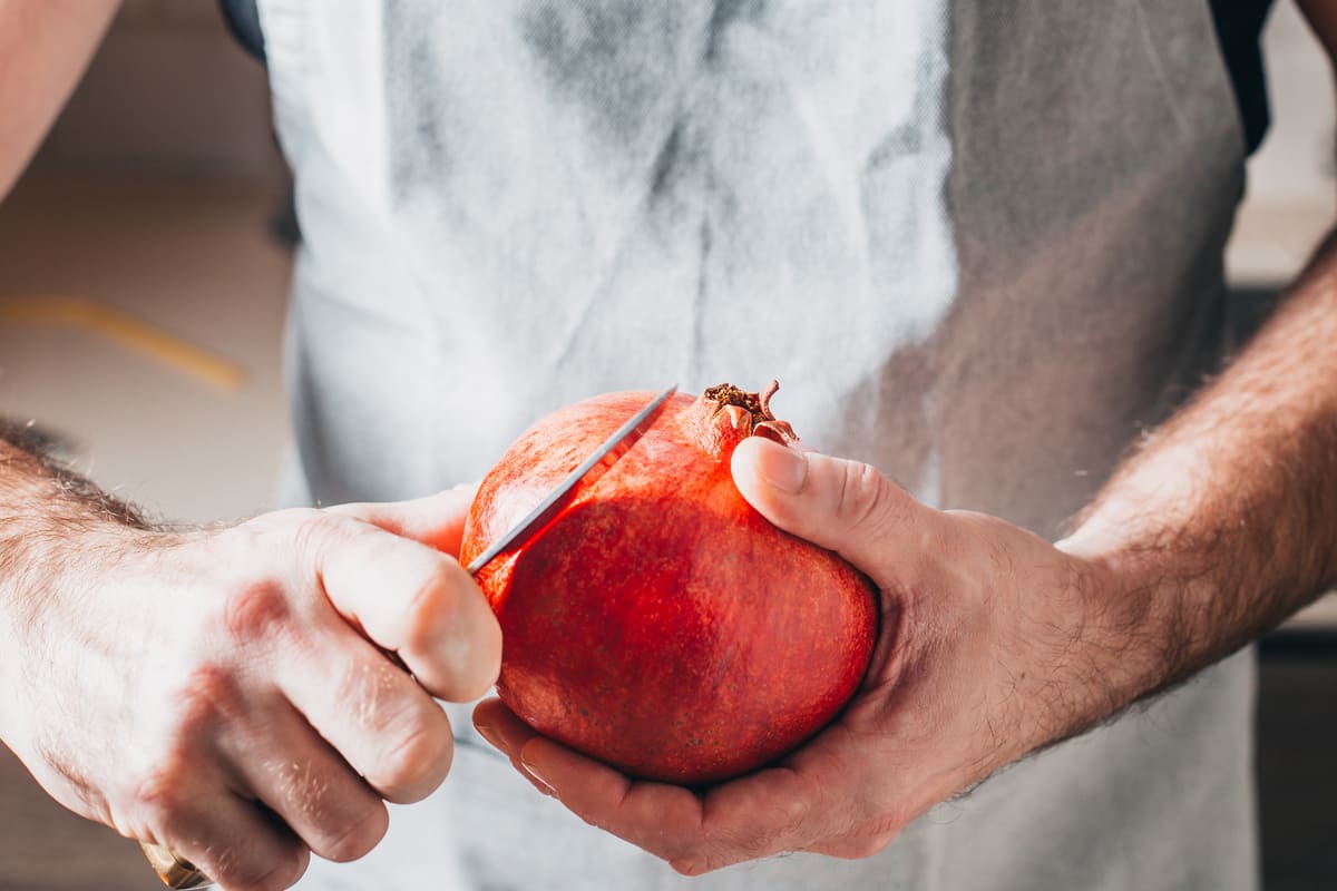 Succo di melograno: la ricetta per farlo in casa, benefici e come conservarlo 1 In primo piano le mani di una persona che incide con un coltello la buccia di una melagrana matura. L’immagine illustra il momento iniziale della preparazione del succo di melograno fatto in casa.
