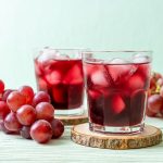 Succo d’uva fatto in casa, bevanda ricca di vitamine e antiossidanti 2 Due bicchieri di succo d’uva rossa con ghiaccio poggiati su sottobicchieri in legno, affiancati da grappoli di uva fresca su un tavolo chiaro.