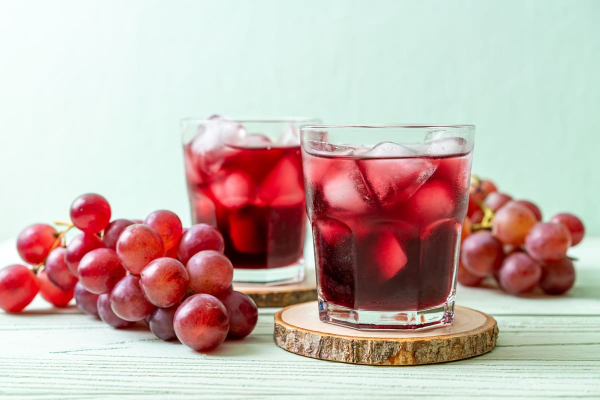 Ricette con uva: 12 idee leggere e gustose 11 Due bicchieri di succo d’uva rossa con ghiaccio poggiati su sottobicchieri in legno, affiancati da grappoli di uva fresca su un tavolo chiaro.