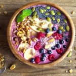 Acai bowl: una ricetta energetica per una colazione sana 1 su tavolo di legno: ciotola di legno con un acai bowl colorata con yogurt greco, semi di chia, bacche di acai, mandorle, frutti di bosco e banane. Accanto cucchiaino con frutta secca