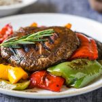 Funghi alla griglia: la bistecca vegetale con il Portobello 1 ricette con funghi: su un piatto bianco in primo piano funghi alla griglia con verdure peperoni rossi e verdi e una foglia di rosmarino