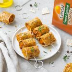 Involtini di zucca e pane azzimo al forno: ricetta e video ricetta di un piatto delizioso 1 su tavolo con tovaglia bianca spiedini di involtini di zucca con accanto la scatola di pane azzimo integrale di Céréal e attorno una bottiglia di olio e altri spiedini