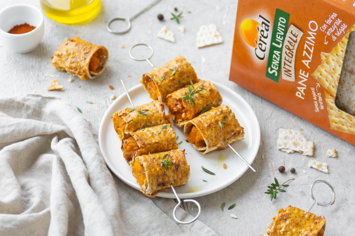 Ricette con la zucca: le migliori idee sane e gustose 35 su tavolo con tovaglia bianca spiedini di involtini di zucca con accanto la scatola di pane azzimo integrale di Céréal e attorno una bottiglia di olio e altri spiedini