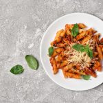 Pasta alla pizzaiola, primo piatto saporito con pomodoro e origano da fare in padella 1 In primo piano un piatto di pasta alla pizzaiola con passata di pomodoro, formaggio grattugiato e foglie di basilico fresco su sfondo grigio.