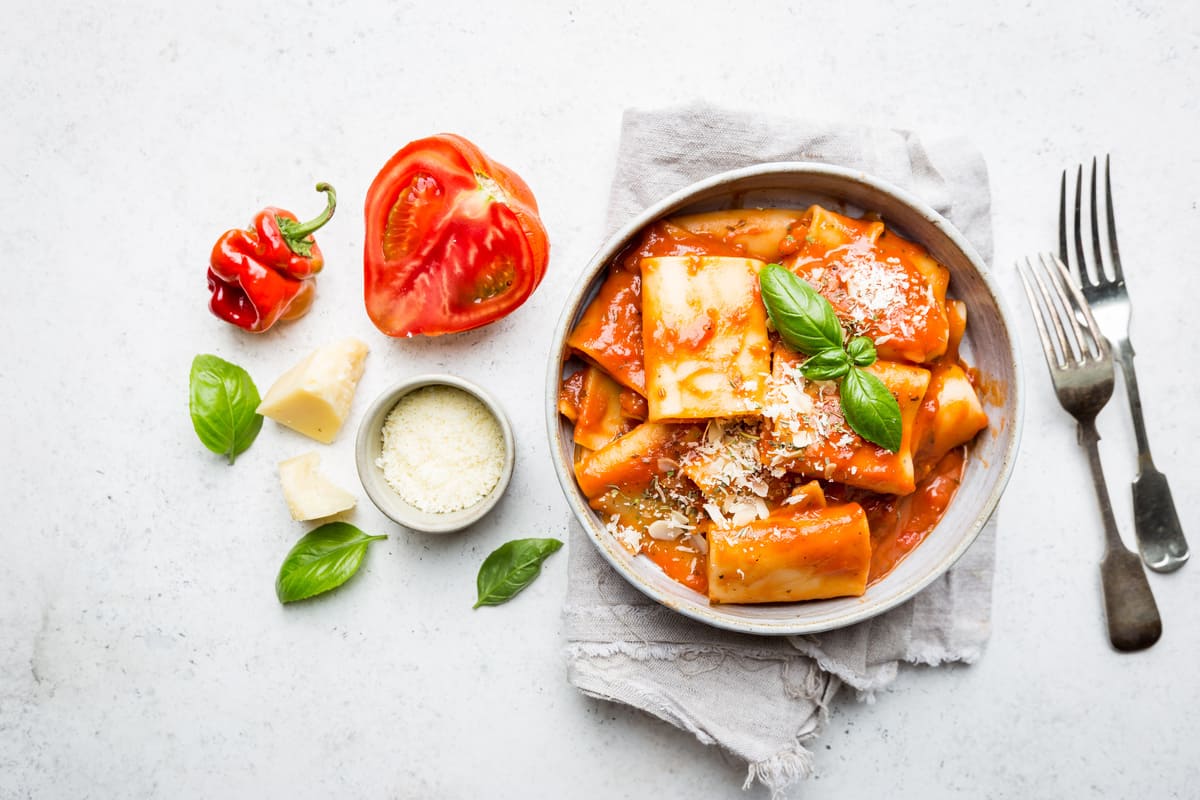 In primo piano un piatto di pasta allo scarpariello con pomodoro, basilico fresco e scaglie di formaggio servito in una ciotola chiara su tovagliolo di lino, con ingredienti freschi come pomodori, peperoncino e formaggio accanto.