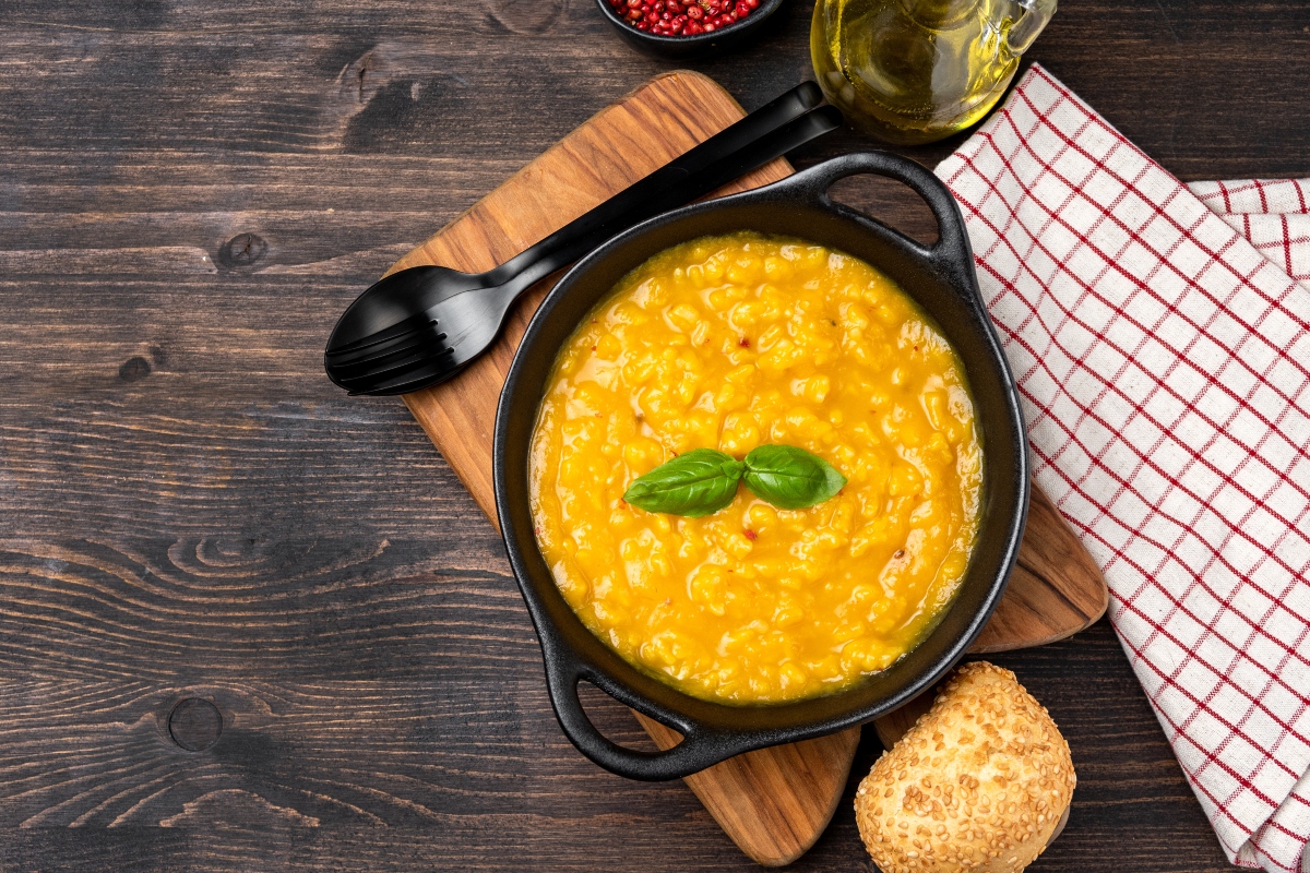 Ricette con la zucca: le migliori idee sane e gustose 9 su tavolo di legno scuro, tagliere di legno chiaro con una ciotola nera piena di pastina con la zucca, accanto cucchiaio nera e dietro canovaccio con a righe bianche e rosse