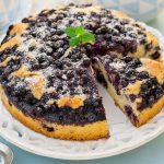Torta ai mirtilli: la ricetta soffice con yogurt, senza burro 2 In primo piano c’è una torta ai mirtilli soffice, decorata con zucchero a velo e servita su un piatto bianco.