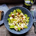 Crema di Pecorino: una salsa gustosa e versatile 2 su tavolo di legno insalata di pollo con la crema di Pecorino in una ciotola blu. Sulla sinistra un pezzo di pecorino toscano DOP su tagliere blu e sulla destra tovagliolo blu con forchetta e coltello. Dietro un bicchiere verde di acqua