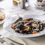 Pasta cozze e pecorino: un primo piatto ricco di sapori 2 su tavolo apparecchiato con tovaglia bianca, piatto fondo con pasta cozze e pecorino, un calice di vino, e un piatto con la focaccia