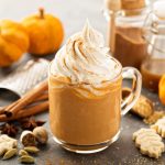 Pumpkin spice latte: la bevanda speziata alla zucca perfetta per l’autunno con videoricetta 1 In primo piano su un tavolo scuro c'è una tazza di vetro con del pumpkin spice latte. Intorno ci sono dei biscotti a forma di foglia, frutta a fuscio e stecche di cannella. Sullo sfondo a sinistra due zucche marroni.