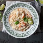 Risotto agli agrumi e gamberi: ricetta di un primo piatto cremoso e delicato 1 In primo piano su un tavolo di legno, c'è un piatto fondo con delle decorazioni verdi, all'interno un cremoso risotto agli agrumi e gamberi con uno spicchio di limone. Sullo sfondo un piattino con degli spicchi di limone a destra, mentre sulla sinistra delle erbette aromatiche.