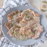 Scaloppine al vino bianco: un secondo piatto di carne leggero e saporito 1 su tavolo, canovaccio e righe bianche e blu sul quale c'è un piatto con scaloppine al vino bianco, con una forchetta sulla destra e un bicchiere di vino bianco e sulla sinistra un cucchiaino con la farina