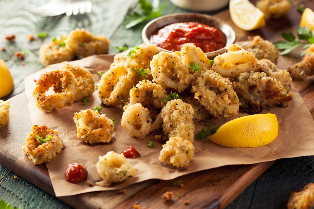 Ricette con friggitrice ad aria semplici e veloci per tutti 1 In primo piano su un tagliere in legno ci sono dei calamari in friggitrice ad aria con delle fettine di limone e una salsa.