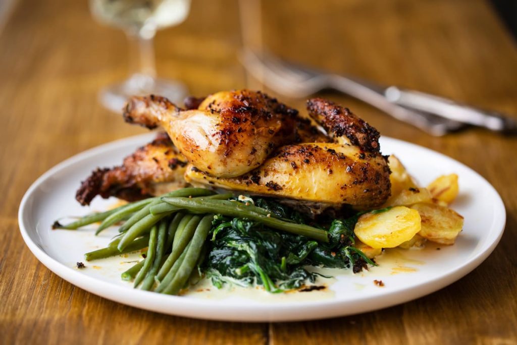 Carne di pollo: che cos'è, valori nutrizionali e calorie dei diversi tagli, benefici, le migliori ricette 11 In primo piano su un tavolo di legno chiaro c'è un piatto bianco con del pollo con friggitrice ad aria e verdure. Sullo sfondo delle posate e un calice sulla sinistra.
