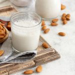 Latte di mandorla fatto in casa: ricetta semplice passo passo 1 In primo piano su un tavolo di marmo chiaro, sulla sinistra c'è un vassoio in legno con un bicchiere di latte di mandorla fatto in casa, accanto un tovagliolo grigio con delle mandorle intorno.