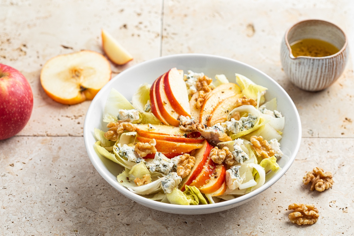 su tavolo da cucina, piatto con indivia belga, gorgonzola e mela rossa tagliata a fette. Attorno contenitore piccolo con olio EVO e la mela tagliata a destra 2 noci