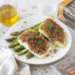Merluzzo in crosta croccante: ricetta con pane di segale e asparagi 1 merluzzo in crosta: piatto bianco tondo con due tranci di merluzzo poggiati su alcuni asparagi, una scatola di pane di segale Céréal accanto