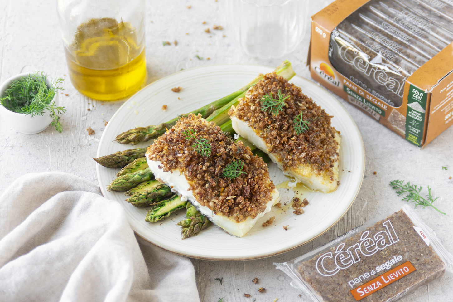 merluzzo in crosta: piatto bianco tondo con due tranci di merluzzo poggiati su alcuni asparagi, una scatola di pane di segale C&eacute;r&eacute;al accanto