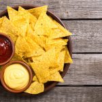 Nachos fatti in casa al forno: croccanti e irresistibili 1 su tavolo di legno un piatto tondo pieno di nachos e due ciotole con salsa rossa e gialla