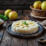 Risotto alle mele: ricetta cremosa e delicata 1 In primo piano su un tavolo di legno c'è un piatto fondo scuro con un risotto alle mele decorato con un fogliolina di menta. Sullo sfondo a sinistra c'è una ciotola con del riso e delle mele in un cestino. In primo piano sulla destra c'è un cucchiaio di legno.