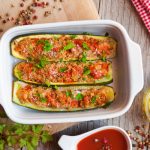 Zucchine ripiene di carne: ricetta facile e gustosa al forno 1 su tavolo di cucina di legno una pirofila bianca con zucchine ripiene al forno e attorno ingredienti come passata di pomodori, olio Evo e pepe