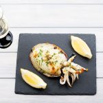 Seppie al limone morbide e saporite: la ricetta con video 1 Su fondo chiaro vassoio piatto nero con seppie al limone e due spicchi di limone.