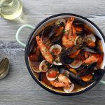 In primo piano su un tavolo da cucina grigio c'è una pentola con la zuppa di cozze alla napoletana. Sulla sinistra delle posate e un piatto fondo giallo.